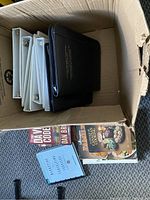 Box with eight white 3-ring binders, three black portfolios, and several books visible including 'The Da Vinci Code' and cookbooks