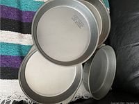 Seven round silver-grey metal cake pans with non-stick surfaces arranged on a black surface with a colorful striped blanket in the background.