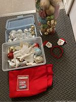 Photo showing two plastic storage bins containing Christmas tree decorations including various baubles and white dove ornaments, with a red stocking and Santa novelty headband on the floor.
