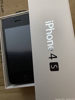 Front view of black Apple iPhone 4S smartphone in original white box, showing screen and home button.
