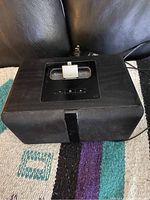 Black speaker dock with Lightning adapter for iPhone, showing volume control buttons on top and fabric-covered front.