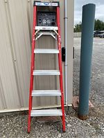 Photo showing a red 6 foot step ladder with aluminum steps against a beige wall outdoors.