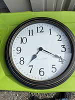 Photo of a large round battery operated wall clock showing white face, black Arabic numerals, black hour and minute hands, and a black frame with gold accent lines. The clock face reads 'Charley Instrument.'