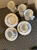 Seven white ceramic cups and seven saucers with a floral pattern and textured surface arranged on a carpeted floor