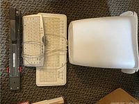 Photo showing a black long reach stapler, regular and travel computer keyboards in beige, and a white plastic room garbage can on carpet.