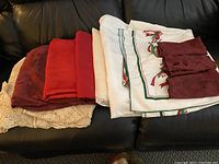 Seven folded table cloths including red, cream crochet, and white holiday-themed cloths, alongside three maroon patterned napkins, all placed on a dark leather couch.