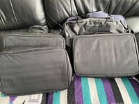 Three black zip-up bags and two larger black soft bags stacked on a leather surface with a colorful rug below.