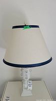 Full view of lamp including cream-colored fabric lampshade with dark blue trim edges and bone china floral decorated base.