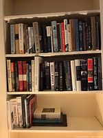 Front view of a bookshelf with three shelves of hardcover and some softcover books, showing bindings and titles.