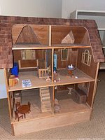 Full view of the handmade wooden doll house showing multiple rooms, wooden flooring, and shingled roof.