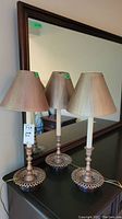 Three matching metal candlestick style table lamps with beige fabric shades on a black surface in front of a large mirror.