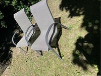 Two matching outdoor chairs with black metal frames and taupe mesh seating placed on grass in a garden, showing the front and side views of the chairs.