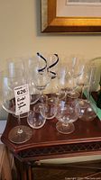 Front view showing seven Riedel wine glasses grouped together with additional glasses and the carafe on a wooden table under a framed picture.