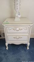 Front view of the off-white bedside table with two drawers and decorative gold handles on a blue carpet.
