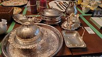 Overview of nine silver plate serving pieces including trays, warmer, pot, basket, and other serving items arranged on a brown table.