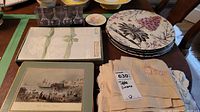 Showcases a table arrangement of boxed napkins, heat resistant mats, decorative plates with floral design, and cream linens with lace.