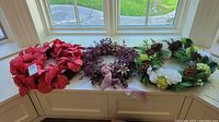 Three faux wreaths displayed together on a white surface in front of a window showing outdoor grassy area. The wreaths include one with large red flowers, one with purple foliage and ribbons, and one with green and white flowers with pinecones.