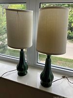 Two side table lamps with blue-green ceramic bases and tall beige fabric shades shown together by window daylight.