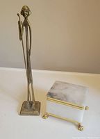 Tall slender brass figurine holding a cane, standing on a square base, shown from front and side angles.