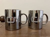 Two shiny metallic mugs with embossed CN logos on a wooden surface.