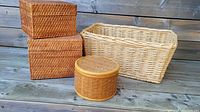 Four wicker baskets: two matching rectangular boxed baskets with lids stacked on one another, large blonde basket with handles, and small round basket with lid in front.