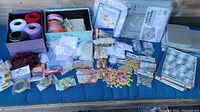 Wide view of assorted crafting supplies laid out on a blue cloth surface including thread, ribbons, bead boards, bells, sequins, and small bags of supplies.