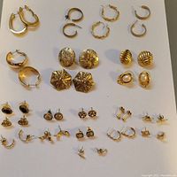 Top-down view of 21 pairs of assorted gold-toned earrings arranged neatly showing a range of sizes and styles including hoops, studs, and decorative buttons.