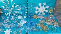 Wide view of various snowflake ornaments in metal, plastic, and beaded materials laid out on blue cushions.
