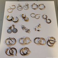 Image showing 15 pairs of silver-toned earrings laid out in rows, various hoop styles, stud and dangling designs, some pairs needing cleaning.