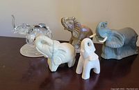Photo showing all five elephant figures on display: Avon cologne bottle, brown stone elephant, white carving, blue-grey figurine, and pink small elephant with chip on ear.