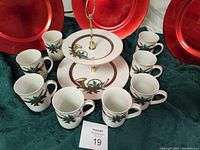 Eight porcelain mugs with holiday greenery design arranged around a two-tiered dessert plate, in original boxes visible.