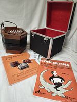 Hexagonal wooden Bonetti Anglo concertina beside the original black hard-sided carrying case with red felt interior and metal latches, two instruction books placed in front