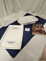 Full lot including Corning Ware serving tray, roasting pan with blue cornflower motif, and cookbook on a dark cloth backdrop.
