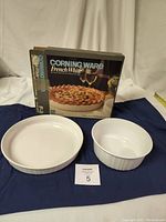 Both CorningWare French White bakeware dishes displayed with original box. One round soufflé dish and one quiche dish shown on a cloth background.