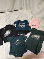 Photo showing five Philadelphia Eagles themed children's clothing pieces and a pink knit hat arranged on a white fabric background.
