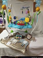 Wide view of Bright Starts Finding Nemo themed baby jumper with attached toys in blue and green, resting on light brown elephant motif blanket with wooden animal decorations and cloth books nearby