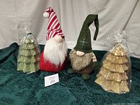 Two Christmas gnomes, one red with a candy cane striped hat and one brown with a green hat, along with two Christmas tree-shaped candles wrapped in plastic, placed on green fabric.