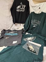 Photo of four apparel items including long sleeve black Super Bowl LII Champions shirt, green Eagles sweatshirt, gray Super Bowl LII Champions shirt, and an Eagles bottle opener and window cling in packaging.