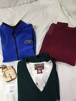 Three clothing items laid out: blue full-zip fleece vest, burgundy long-sleeved pullover sweater, and green sleeveless knit vest with V-neck, along with a small wooden golf bag picture frame and lot number card