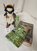 Linnea cloth doll seated in wooden rocking chair with attached small storybook, two additional books arranged in front.
