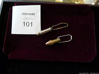 Two metal bullet casing replica keychains, one silver-tone and one brass-tone on a dark maroon surface with auction number 101.