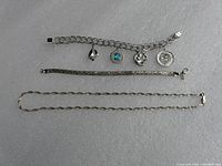 Three sterling silver jewelry pieces arranged flat on a gray surface including a charm bracelet with four charms, a narrow herringbone bracelet, and an elongated link necklace.