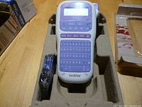 Brother P-Touch handheld ribbon label maker, purple and white with QWERTY keyboard and LCD display, placed in molded cardboard packaging alongside a ribbon cartridge.