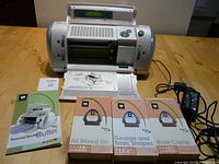 Full view of Cricut machine on table with power cord, user manual, and three font cartridge sets displayed in front.