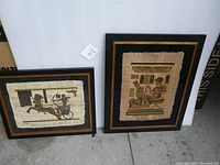 Photo of two framed Egyptian art prints on papyrus resting against a white background. Left print depicts horse and chariot, right print depicts seated figure.