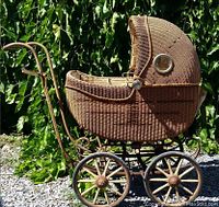 Side view of antique wicker baby carriage showing woven wicker body, metal handle, and wooden wheels with rubber.