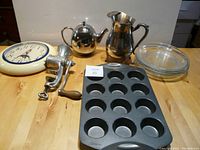 Photo showing the entire lot: 4 Pyrex pie plates stacked, silver-plated jug, Epicure teapot, Wilton muffin tin, vintage food grinder, and Ingram wall clock.