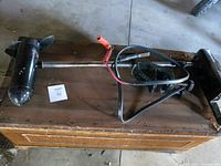 Black Motor Guide Stealth 2000V trolling motor laid horizontally on wooden surface showing motor and clamp base with attached cables and clips.