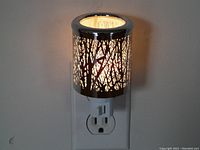 Side view of the nightlight plugged into wall outlet showing the silver etched tree branch pattern illuminated from within.