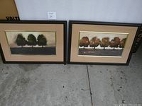 Pair of framed prints placed side by side on the floor, showing two different stylized tree scenes in dark wood frames with beige matting and gold inner trim.